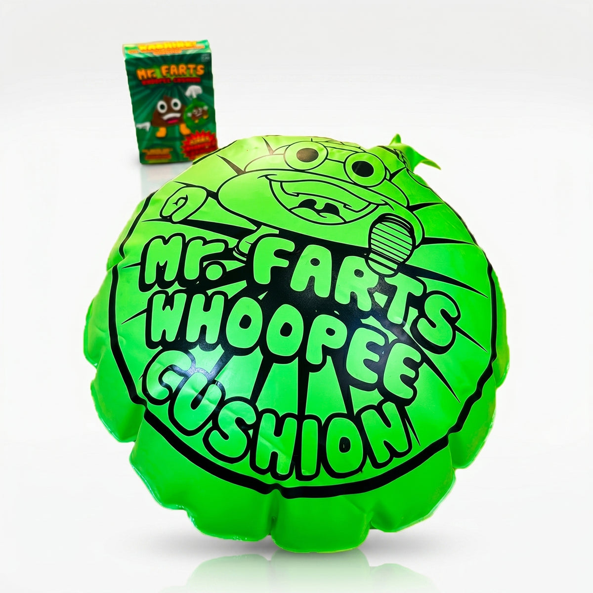 A giant green Mr farts Whoopee cushion centred on a clean background with a Mr Farts Giant Whoopee Cushion packaging slightly blurred behind it.