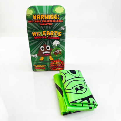 A giant green Mr farts Whoopee cushion folded up, recently out of it's packaging on a clean background with a Mr Farts Giant Whoopee Cushion packaging box open behind it. 