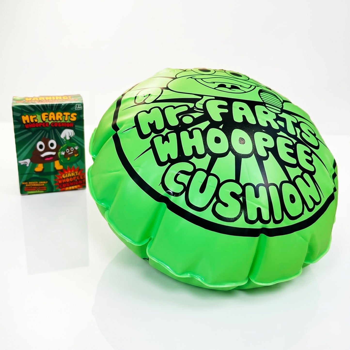 A giant green Mr farts Whoopee cushion on a clean background with a Mr Farts Giant Whoopee Cushion packaging slightly blurred behind it. 